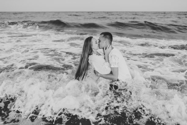 游泳。穿着湿衣服的男人和女人在海里。一对情侣在海边拥抱亲吻。男性亲吻和拥抱女性站在水与大浪的海洋，享受夏日。黑白照片图片下载