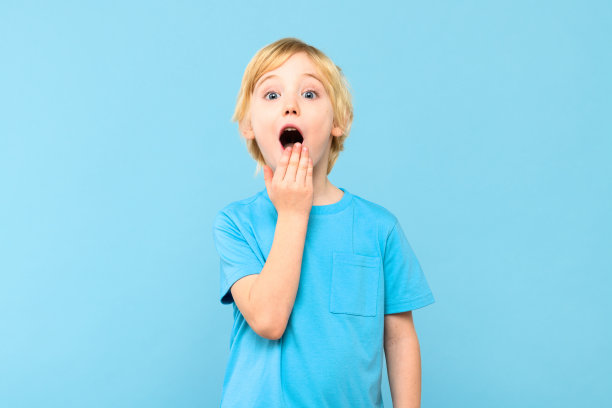 哇!一个震惊的可爱小男孩的肖像，金色的头发用一只手遮住张开的嘴，在柔和的蓝色背景上。惊讶的学龄前儿童摄影棚照片。图片下载