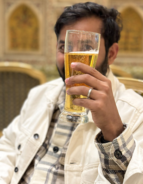 印度男子用威森酒杯喝酒的特写照片，啤酒上的气泡形成了泡沫的泡沫头，抬高的酒杯在脸前，酒吧作为背景，焦点在前景图片下载