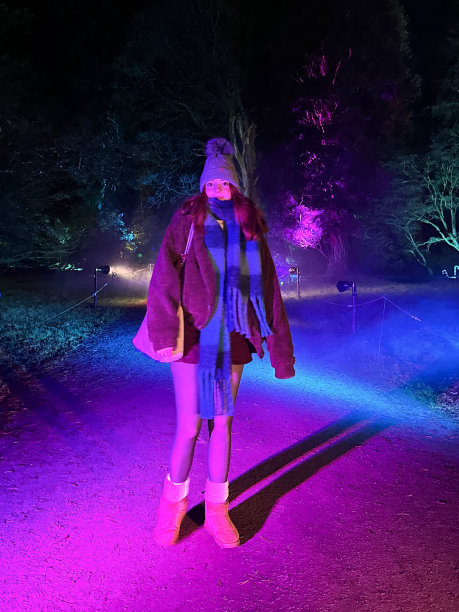 特写图片，红头发的女人戴着羊毛球帽，围巾和羊毛夹克走在照亮林地小径在植物园，背着肩袋，圣诞灯，黑暗，夜空，重点在前景图片下载