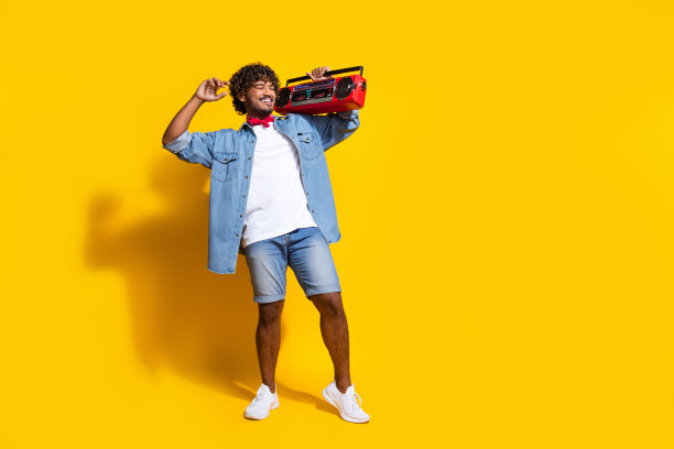 漂亮的年轻男性的全尺寸照片拿着立体声音箱迪斯科舞厅穿着时尚的牛仔服装红色围巾孤立的黄色背景图片下载