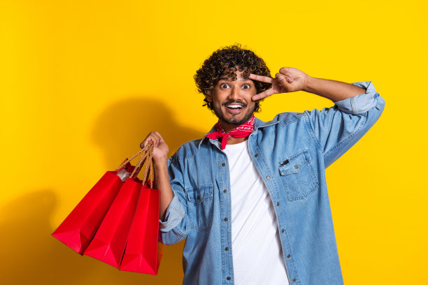 照片中，帅气的年轻人购物时展示v字脸，穿着时髦的牛仔服装，红色围巾孤立在黄色背景上图片下载