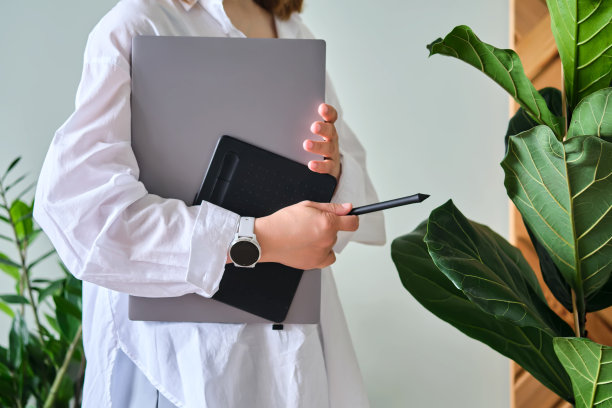 一个女人拿着笔记本电脑和图形平板电脑，背景是绿色植物。图片下载