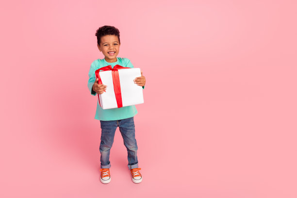 迷人的小男孩拿着礼盒走路的全尺寸照片，穿着时髦的海蓝宝石服装，孤立在粉红色的背景上图片下载