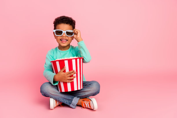 可爱小男孩的全身照片吃爆米花桶坐在地板上穿着时髦的海蓝宝石服装孤立在粉红色的背景图片下载