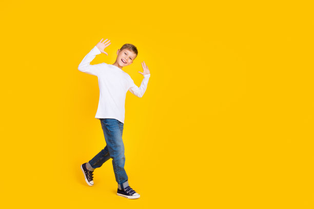 友好时髦的孩子穿着白衬衫，挥舞着手臂，他走空空间孤立的黄色背景的全长照片图片下载