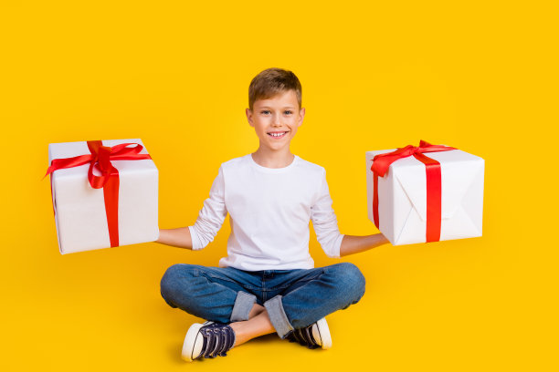 正面开朗的孩子穿着白衬衫的全长照片比较两个礼物盒孤立的黄色背景图片下载