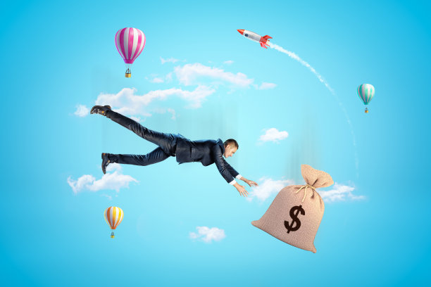 年轻的商人试图抓住帆布钱袋，在蓝色的天空中坠落，背景是热气球和火箭。图片下载
