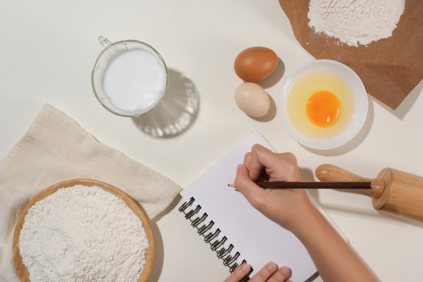 烹饪概念照片，一位女性手拿着铅笔在笔记本上写菜谱，周围是一些烘焙糕点的配料。用于设计或显示元素的空间图片下载