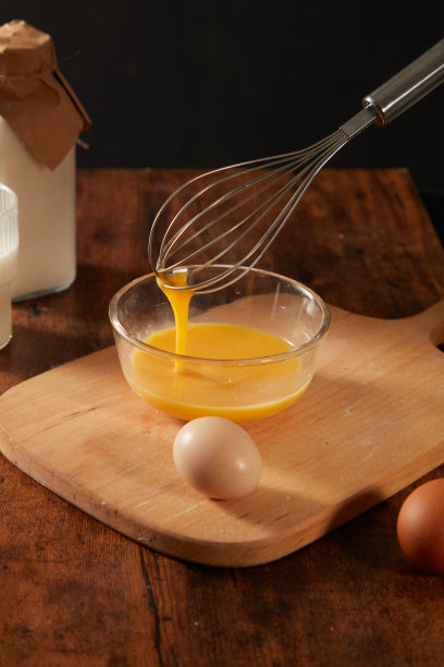 搅蛋器在透明的玻璃碗里搅拌鸡蛋，放在木制案板上，旁边是木桌上自制烘焙的新鲜鸡蛋。正面照片图片下载