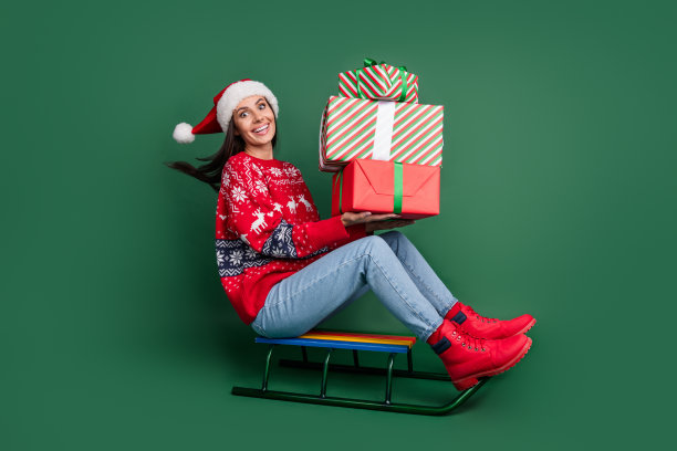 圣诞老人的全身照片帮助小精灵年轻的女士骑雪橇拿着一堆礼盒丑陋的毛衣孤立的绿色背景图片下载