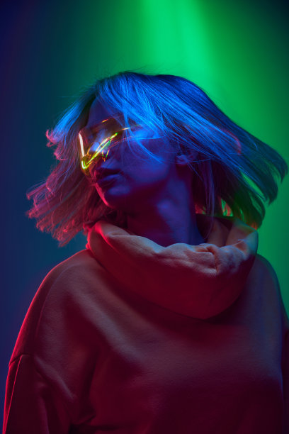 舒适与高科技时尚的结合。年轻的女孩戴着全息眼镜和休闲帽衫，在霓虹灯下渐变绿蓝色背景图片下载