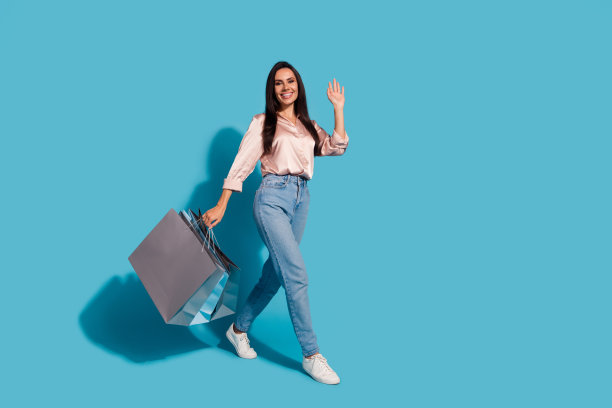 开朗的女孩全身照片穿着丝绸衬衫牛仔裤拿着购物袋走空空间说hi孤立在蓝色的背景图片下载