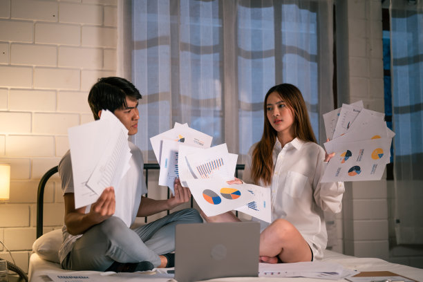 抑郁的亚洲年轻夫妇在漆黑的夜晚过度工作。迷人美丽的商人和女人坐在床上，在家里手里拿着许多文件，感到担心和沮丧。图片下载