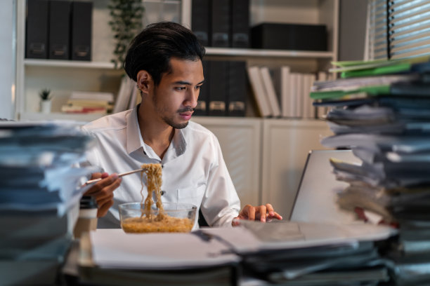 亚洲年轻商人晚上在办公室工作时吃面条。有吸引力的职业男性员工坐在桌子上，感到饥饿和饥饿，然后在公司工作场所吃晚餐。图片下载