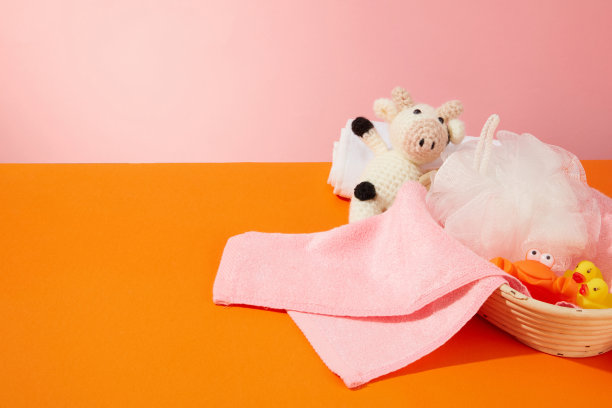 一个白色的婴儿沐浴产品篮子，里面有毛巾、玩具和沐浴海绵，在橙色桌子的左边，粉红色的背景。前视图照片与空白空间显示产品图片下载
