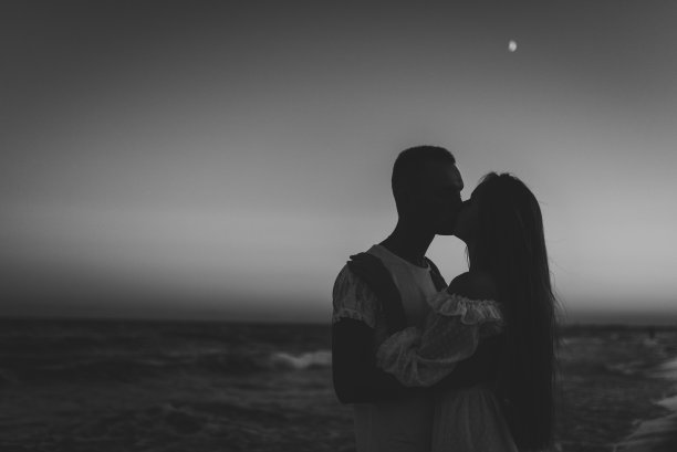 夕阳下男人亲吻女人的剪影，背景是天空中的月亮。女人亲吻男人，享受夏日的黄昏。一对在沙滩上相爱的情侣。放文字的地方。黑白照片图片下载