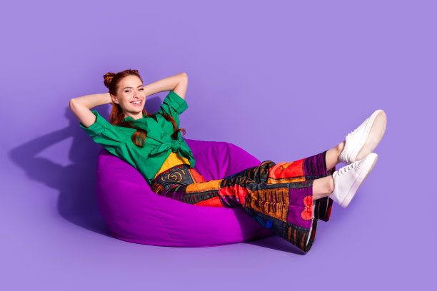 长照片的好心情的女孩穿着印花复古裤坐在pouf保持手臂背后的头冷孤立在紫罗兰色的背景图片下载
