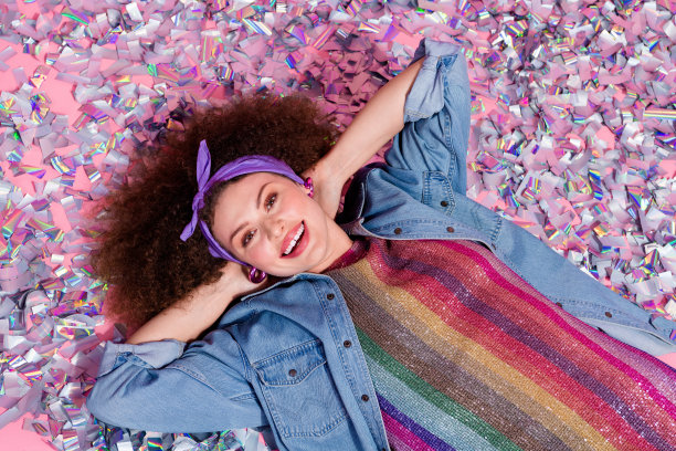 照片中可爱漂亮的女士穿着牛仔裤，手臂放在脑后，躺着五彩纸屑孤立的粉红色背景图片下载