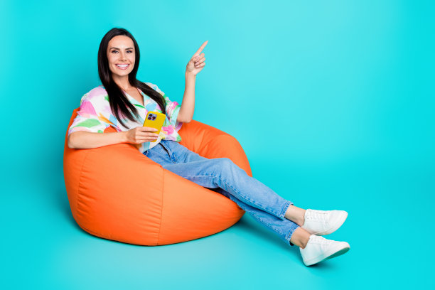 照片中开朗的女人穿着时尚的印花衣服，坐在舒适的椅子上，推荐手机，在青色背景上孤立的空间图片下载