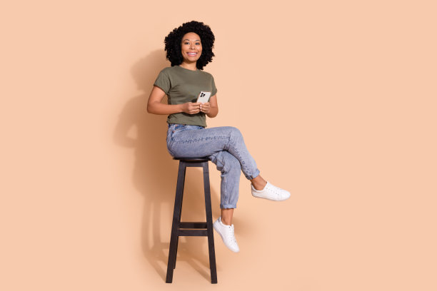 迷人的学生全身照片，穿着卡其色t恤牛仔裤，拿着智能手机，坐在酒吧凳子上，孤立的米色背景图片下载