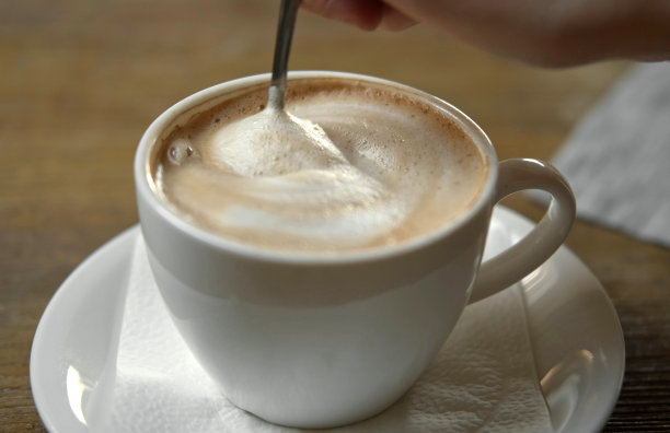 手正在搅拌拿铁的奶泡。用钢勺搅拌卡布奇诺。热咖啡在白色经典陶瓷杯在木桌。现代咖啡店，自助餐厅。厚的泡沫。特写镜头图片下载