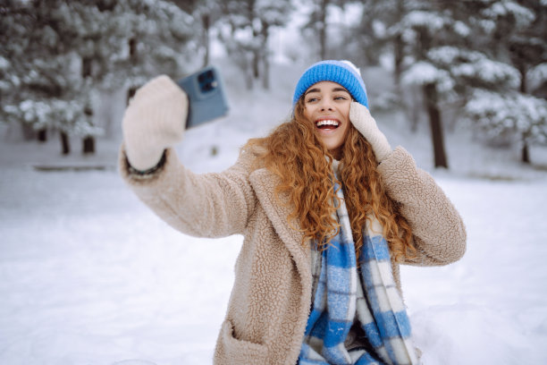 一个快乐的女人在下雪的冬季森林里用智能手机自拍。图片下载