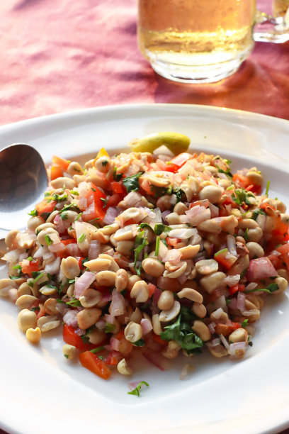 特写图片白色碗的花生masala chaat，街头风味小吃，花生，chaat masala，辣椒粉，盐，柠檬汁香菜，切片serrano胡椒(辣椒)，番茄丁和红洋葱，金属勺子，冷凝在玻璃酒杯上图片下载