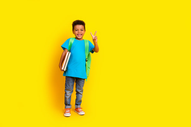 全尺寸照片，快乐的男孩穿着休闲的蓝色t恤牛仔裤帆布背包显示v字抱书孤立在生动的黄色背景图片下载