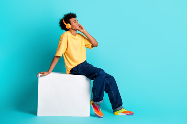 全尺寸照片，无忧无虑的男人穿着超大号的t恤牛仔裤，坐在站台上，在蓝绿色的背景下，戴着耳机欣赏音乐图片下载