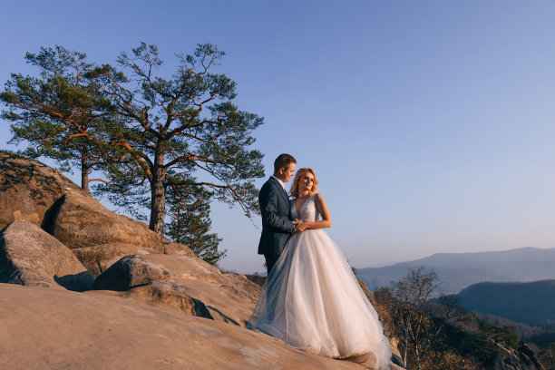 这是一对浪漫的婚礼夫妇的美丽照片，新娘和新郎穿着华丽的婚纱，拥抱在山上的大岩石旁。图片下载