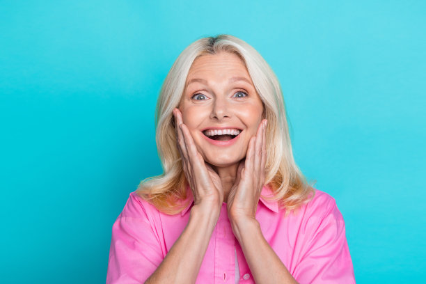 照片中，迷人的退休妇女抚摸着脸颊，震惊地穿着时尚的粉色智能休闲服装，背景是海蓝宝石色图片下载