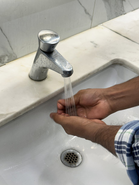 特写照片:一个无法辨认的人站在公共浴室的水池边，在自来水下洗手，水龙头在流动，白色大理石浴室柜台在高处图片下载