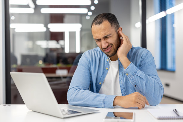 一位在办公室工作的西班牙裔年轻人，坐在办公桌前，把手放在耳朵上方，感到剧烈的疼痛和充血图片下载