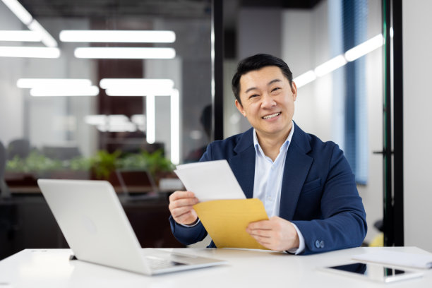 兴奋的日本商人在阅读了文件中令人愉快的信息后，对着镜头开心地微笑。满意的男性在台式机和电脑上工作，并批准信贷申请图片下载