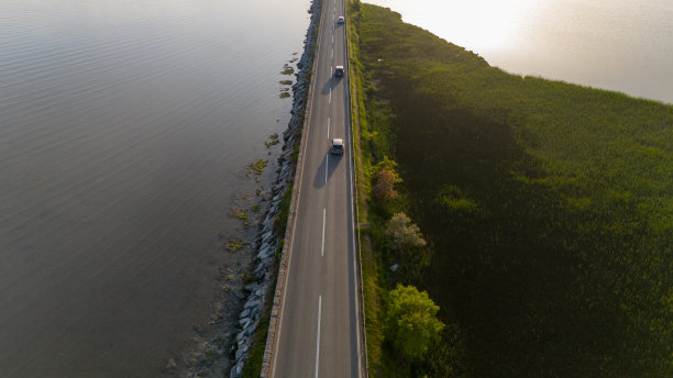 日落时分，两辆汽车行驶在海湾对面的公路上的航拍照片图片下载