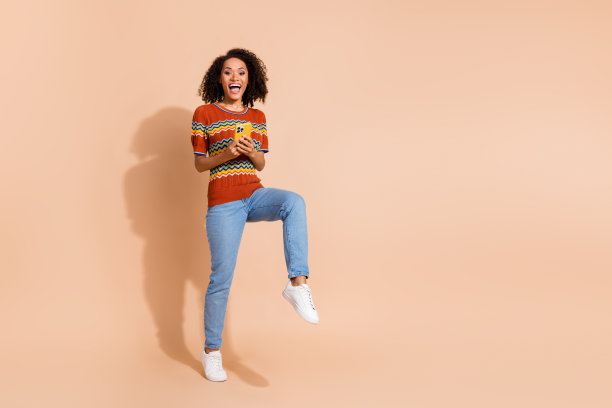 全尺寸照片积极的女人穿针织t恤牛仔裤拿智能手机站在一条腿上孤立的米色背景图片下载