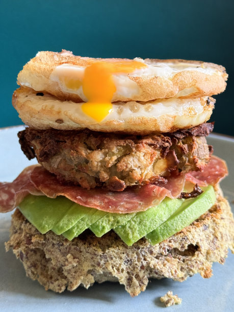 自制早餐汉堡堆的特写图片，白色盘子上的全麦面包上放着牛油果片、意大利腊肠、土豆泥和两个煎蛋，黄色的蛋黄，蓝绿色的背景，重点放在前景上图片下载
