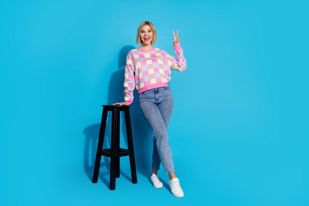 乐观的女孩穿着格子套头衫的全长照片，手放在酒吧椅子上，蓝色背景上显示v字符号图片下载