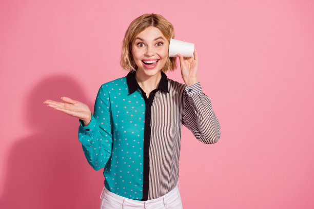 漂亮的年轻女孩的照片肖像听纸杯说话电话穿着时髦的条纹青色服装孤立在粉红色的背景图片下载