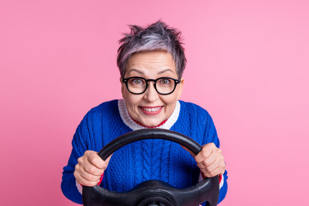 开朗有趣的退休妇女的照片，穿着时髦的蓝色衣服，拿着方向盘，驾驶自己的车，孤立在粉红色的背景上图片下载