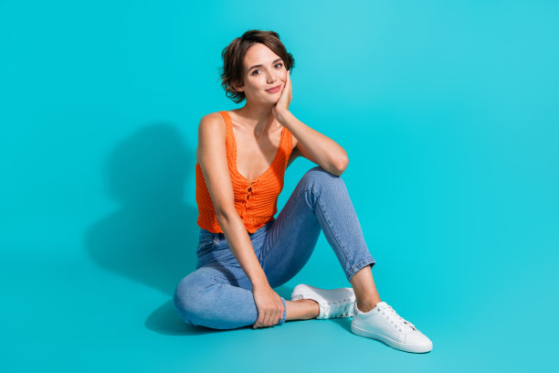 全身照片可爱的积极的女人穿着橙色的牛仔裤坐在地板上，手掌在脸颊上孤立的绿松石色背景图片下载