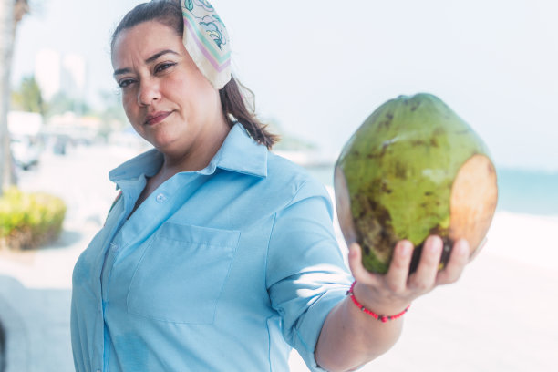 一个拉丁女人在海滩上喝椰子汁，在街边的小摊上拿着几杯热带水果。图片下载
