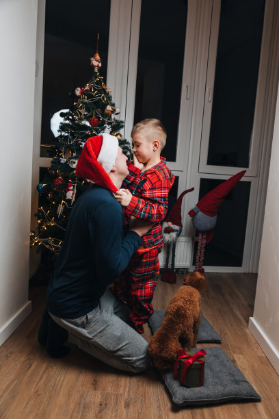 穿着红色睡衣的父子带着狗坐在圣诞树旁拥抱。前视图图片下载