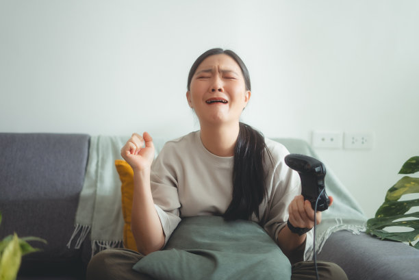 亚洲女子用操纵杆玩游戏的正面照片，输了游戏后坐在家里的沙发上哭泣，感到沮丧。图片下载