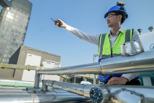在晴朗的天空下，戴着安全帽和安全背心的工程师在建筑工地用对讲机与城市建筑进行通信。图片下载