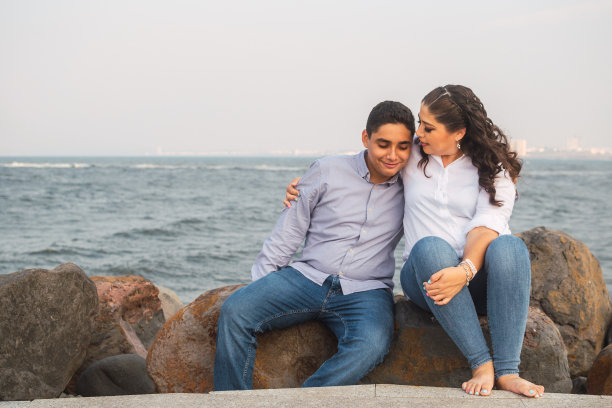 母亲节期间，一位年轻的拉丁妇女抱着她十几岁的儿子坐在海边的岩石上图片下载