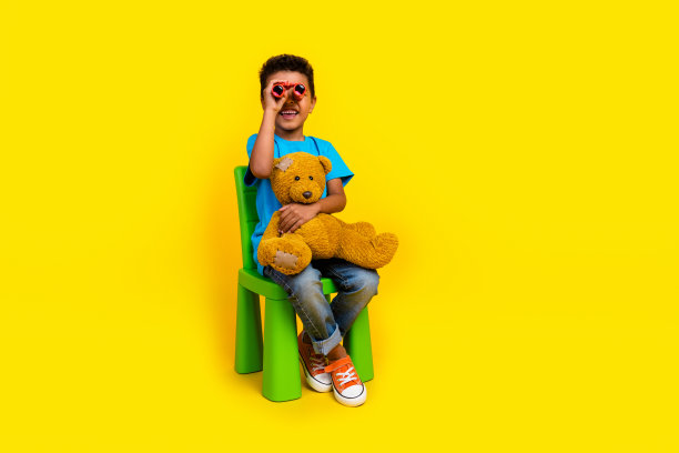 穿着蓝色t恤的时髦小男孩的全身照片，在充满活力的黄色背景下，拿着泰迪熊看双筒望远镜图片下载