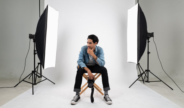 年轻聪明的亚洲专业摄影师的肖像照片坐在白色的照片背景室内摄影棚与一套频闪闪光灯和柔光箱准备产品和模型拍摄。图片下载