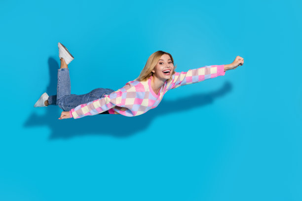 可爱的女孩穿着彩色套头衫在空旷的空间里飞行，就像超级女人被隔离在蓝色的背景上图片下载